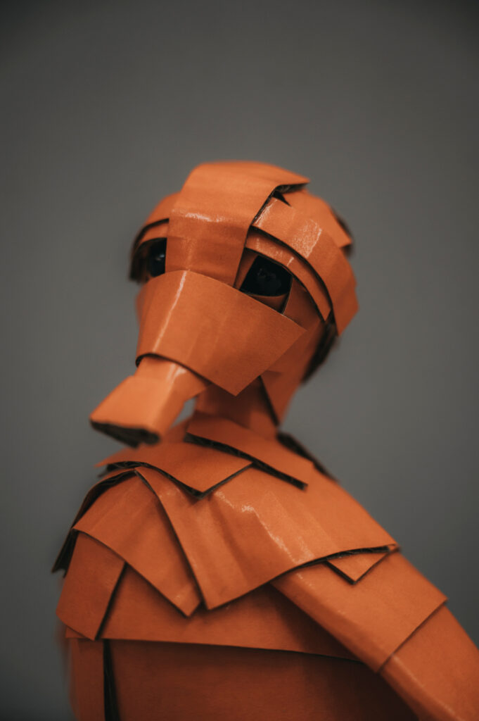 Patito naranja sobre fondo gris. Escultura de cartón. Orgaz