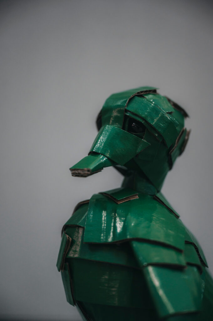 Patito verde inglés de perfil, fondo gris. Escultura de cartón. Orgaz